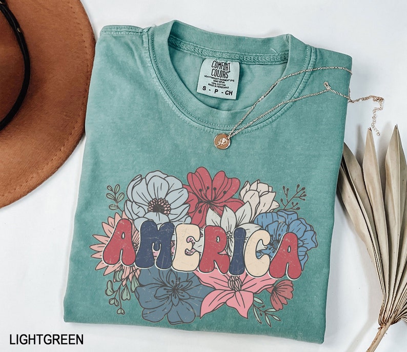 May include: Light green t-shirt with a floral design and the word "AMERICA" in colorful, retro-style lettering. The shirt is made of a soft, comfortable material and has a relaxed fit.