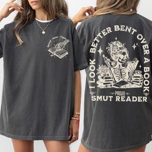 May include: A dark gray oversized t-shirt with a skeleton reading a book graphic and the text "I look better bent over a book, Proud Smut Reader".