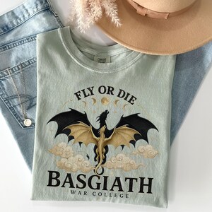 May include: A pale green t-shirt featuring a dragon design with the text "FLY OR DIE" and "BASGIATH WAR COLLEGE". The dragon has black wings and a gold body, with clouds and moon phases. The shirt is folded on top of blue denim jeans.