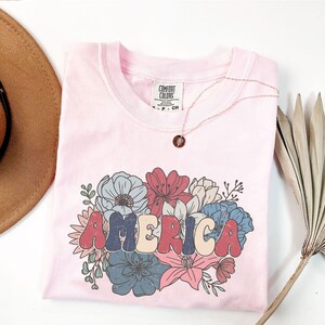 May include: A pink t-shirt with a floral design and the word "AMERICA" in colorful, retro-style lettering. The shirt has a tag that reads "Comfort Colors".