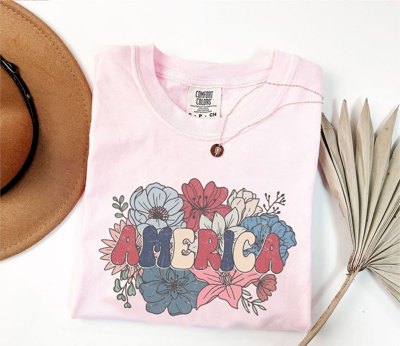 May include: A pink t-shirt with a floral design and the word "AMERICA" in colorful, retro-style lettering. The shirt has a tag that reads "Comfort Colors".