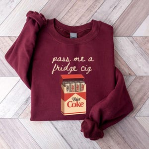 May include: Burgundy sweatshirt with the phrase "pass me a fridge cig" in a cursive font. Below the text is a graphic of a Diet Coke cigarette pack with Diet Coke cans inside.