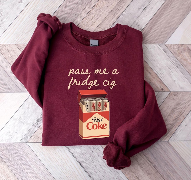 May include: Burgundy sweatshirt with the phrase "pass me a fridge cig" in a cursive font. Below the text is a graphic of a Diet Coke cigarette pack with Diet Coke cans inside.