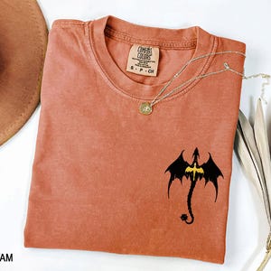 May include: A rust-colored t-shirt with a black dragon graphic and a gold chain necklace. The shirt has the text "COMFORT COLORS" and "S - P - CH" printed on the tag.