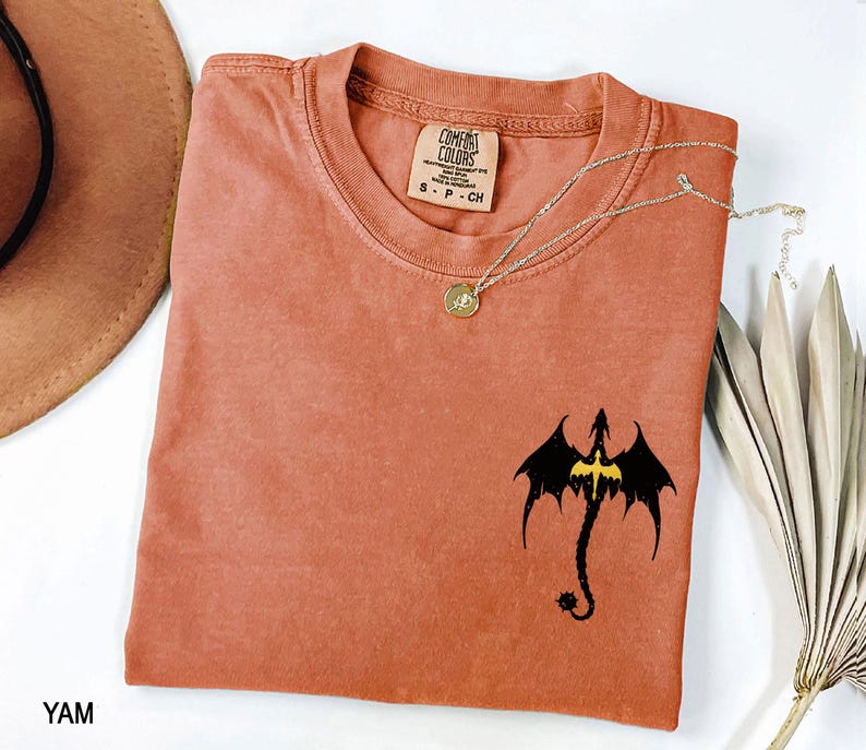 May include: A rust-colored t-shirt with a black dragon graphic and a gold chain necklace. The shirt has the text "COMFORT COLORS" and "S - P - CH" printed on the tag.