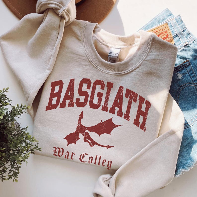 May include: A beige sweatshirt with the word "BASGIATH" in distressed red lettering, along with a red dragon graphic and the words "War Colleg". The sweatshirt is folded, with a portion of a brown hat and denim shorts visible.