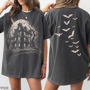 May include: Dark gray t-shirt featuring a haunted house graphic on the front and a bat design on the back. The front graphic shows a spooky house with a full moon and bare trees. The back of the shirt has a flock of bats in flight.