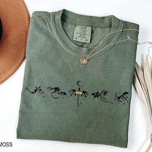 May include: Moss green t-shirt with a crew neck and a black dragon graphic. The shirt has a small Comfort Colors tag and a gold necklace with a pendant. The word "MOSS" is printed at the bottom left.