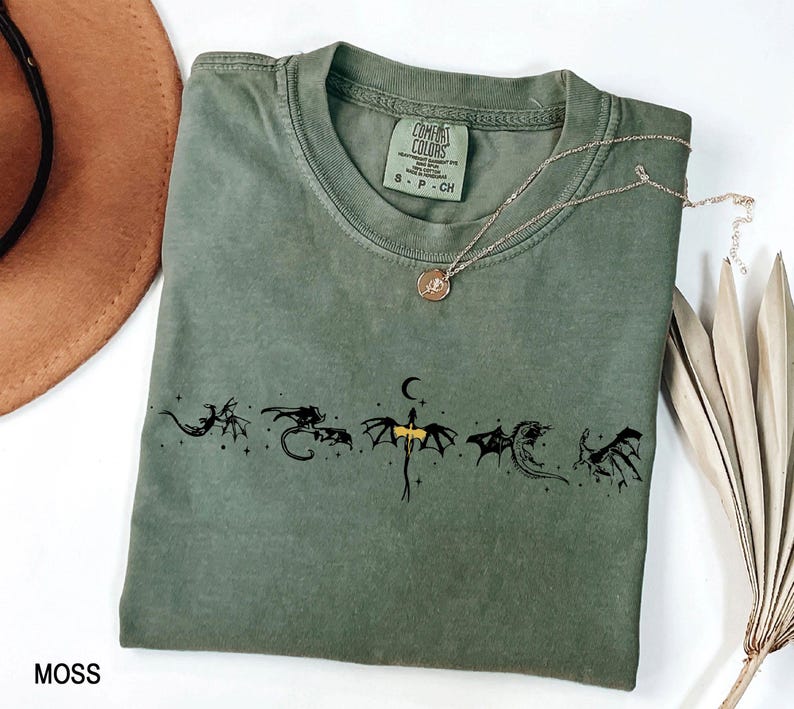 May include: Moss green t-shirt with a crew neck and a black dragon graphic. The shirt has a small Comfort Colors tag and a gold necklace with a pendant. The word "MOSS" is printed at the bottom left.