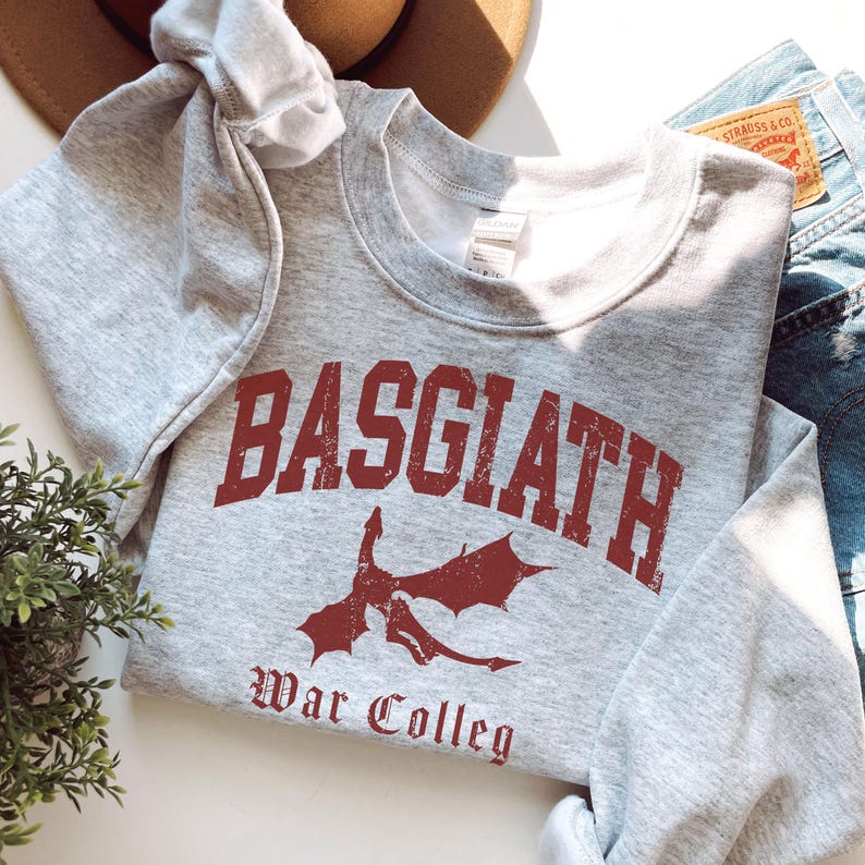 May include: A heather gray sweatshirt with the word "BASGIATH" in distressed maroon lettering. Below the word is a maroon dragon graphic and the words "War Colleg". The sweatshirt is folded on a white surface with a pair of blue jeans and a brown hat.