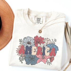 May include: A beige t-shirt with a floral design and the word "AMERICA" in colorful, retro-style lettering. The shirt has a tag that reads "COMFORT COLORS" and size information.