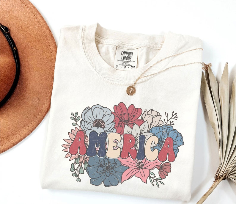 May include: A beige t-shirt with a floral design and the word "AMERICA" in colorful, retro-style lettering. The shirt has a tag that reads "COMFORT COLORS" and size information.