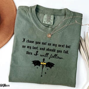 May include: An olive green t-shirt with the text "I chose you not as my next but as my last, and should you fall, then I will follow." A black and yellow dragon graphic is below the text. A gold necklace and a brown hat are also in the image.