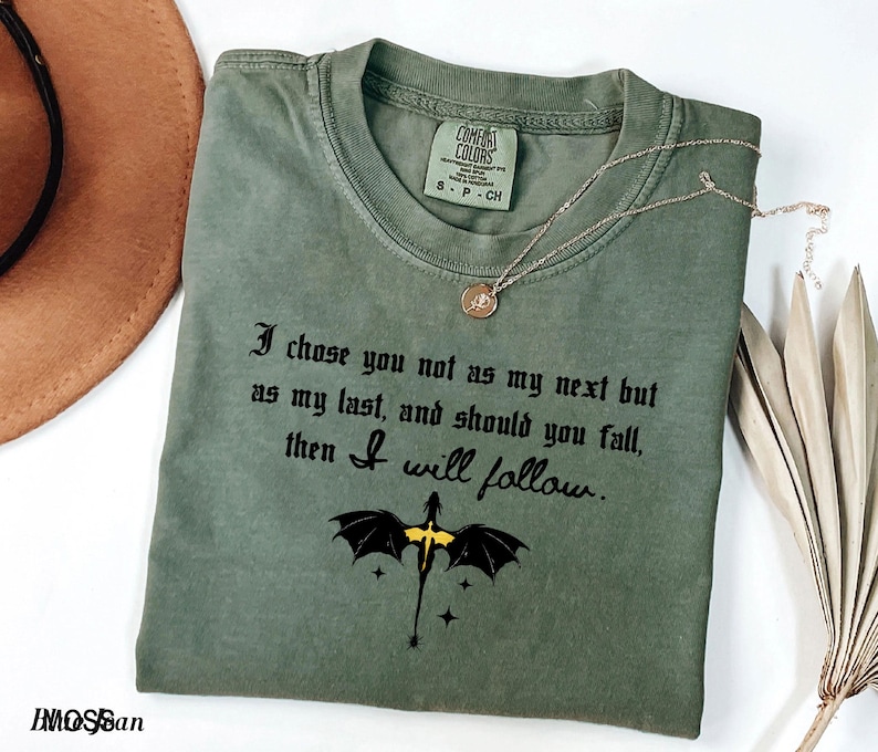 May include: An olive green t-shirt with the text "I chose you not as my next but as my last, and should you fall, then I will follow." A black and yellow dragon graphic is below the text. A gold necklace and a brown hat are also in the image.