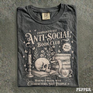 May include: Dark gray t-shirt with the text "UNAPOLOGETICALLY ANTI-SOCIAL BOOK CLUB" and a graphic of a skull, books, and a teacup. The shirt also has the text "MAKING FRIENDS WITH CHARACTERS, NOT PEOPLE".