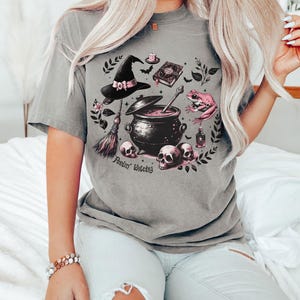 May include: A gray t-shirt with a witchy graphic featuring a cauldron, a witch's hat, a frog, a broom, and skulls. The text "Feelin' Witchy" is printed on the shirt.