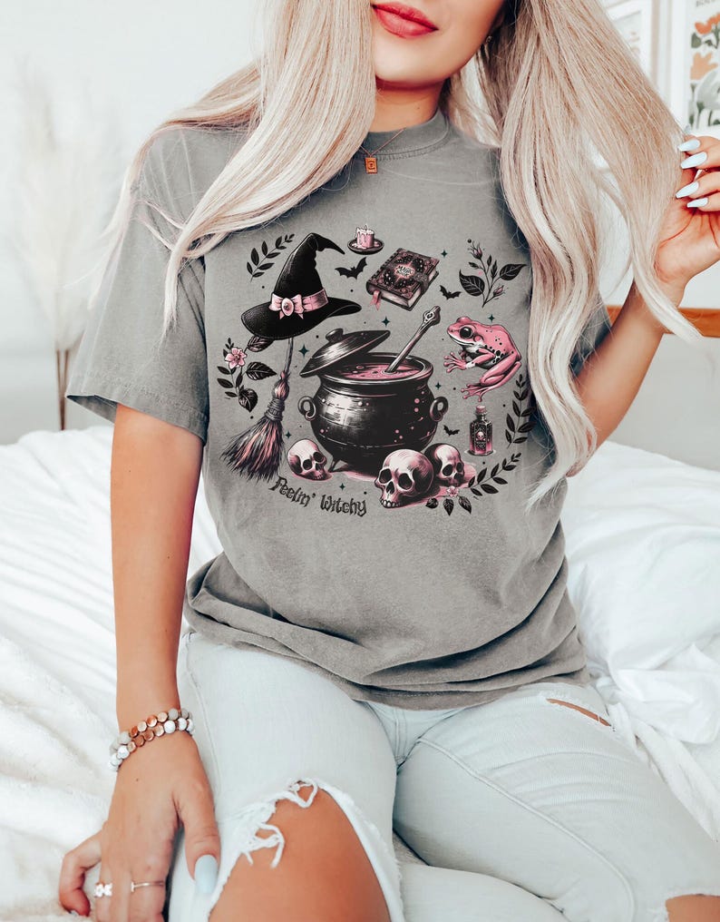 May include: A gray t-shirt with a witchy graphic featuring a cauldron, a witch's hat, a frog, a broom, and skulls. The text "Feelin' Witchy" is printed on the shirt.