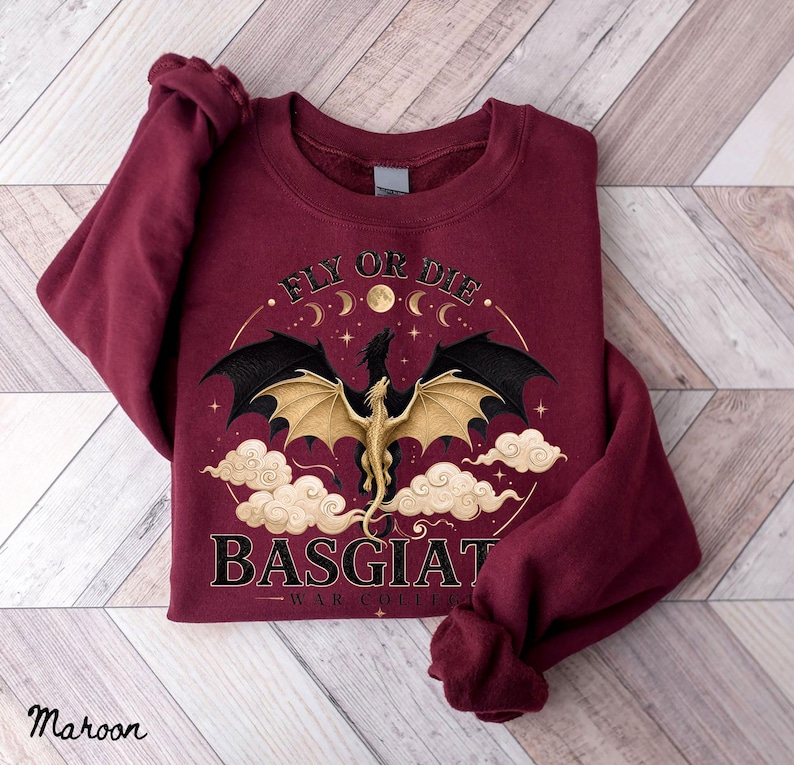 May include: Maroon sweatshirt with a dragon graphic. The design features a gold dragon with black wings, clouds, and the text "FLY OR DIE" and "BASGIAT - WAR COLLECTION". The sweatshirt is a solid maroon color.