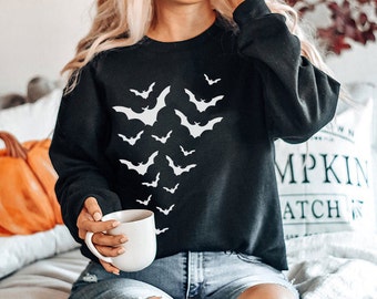 May include: A black sweatshirt with a white graphic print of bats. The sweatshirt is worn by a person holding a white mug.