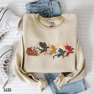 May include: A beige crewneck sweatshirt featuring a graphic print of Santa Claus in a sleigh pulled by four flying dragons in black, gold, blue, and red. The design includes a full moon and small stars. The sweatshirt is layered over blue denim jeans.