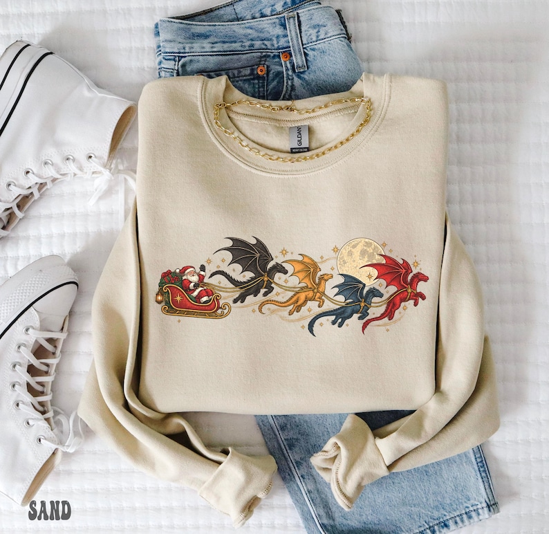 May include: A beige crewneck sweatshirt featuring a graphic print of Santa Claus in a sleigh pulled by four flying dragons in black, gold, blue, and red. The design includes a full moon and small stars. The sweatshirt is layered over blue denim jeans.