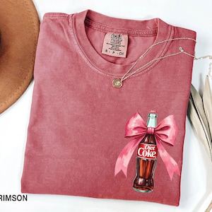 May include: Crimson red t-shirt with a watercolor illustration of a Diet Coke bottle with a pink bow. The shirt has a Comfort Colors tag with the size S-P-CH.