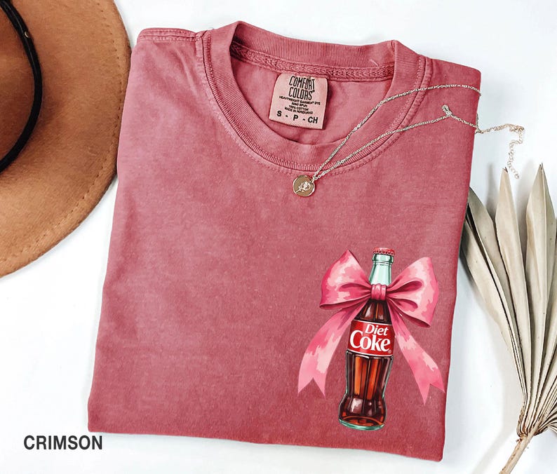 May include: Crimson red t-shirt with a watercolor illustration of a Diet Coke bottle with a pink bow. The shirt has a Comfort Colors tag with the size S-P-CH.
