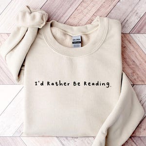 K&ouml;nnte beinhalten: Ein beiger Pullover mit dem Aufdruck "I'd Rather Be Reading." in Schwarz.