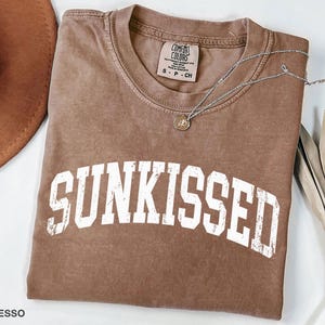 May include: Espresso-colored t-shirt with the word "SUNKISSED" in white, distressed lettering. A silver necklace with a small pendant rests on the shirt. The shirt is folded, and a brown hat is partially visible in the upper left corner.