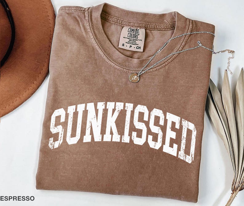 May include: Espresso-colored t-shirt with the word "SUNKISSED" in white, distressed lettering. A silver necklace with a small pendant rests on the shirt. The shirt is folded, and a brown hat is partially visible in the upper left corner.