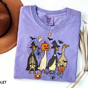 Puede incluir: Una camiseta violeta Comfort Colors con un dise&ntilde;o de Halloween. El gr&aacute;fico muestra cuatro patos de dibujos animados con disfraces de Halloween, como un esqueleto, una calabaza y una bruja. La camiseta tambi&eacute;n tiene una telara&ntilde;a, murci&eacute;lagos y fantasmas.