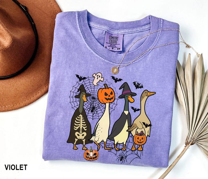 Puede incluir: Una camiseta violeta Comfort Colors con un dise&ntilde;o de Halloween. El gr&aacute;fico muestra cuatro patos de dibujos animados con disfraces de Halloween, como un esqueleto, una calabaza y una bruja. La camiseta tambi&eacute;n tiene una telara&ntilde;a, murci&eacute;lagos y fantasmas.