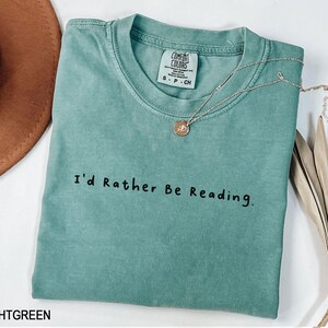 May include: Light green t-shirt with the text "I'd Rather Be Reading." printed in black. The shirt has a distressed look and is made of a soft, comfortable fabric.