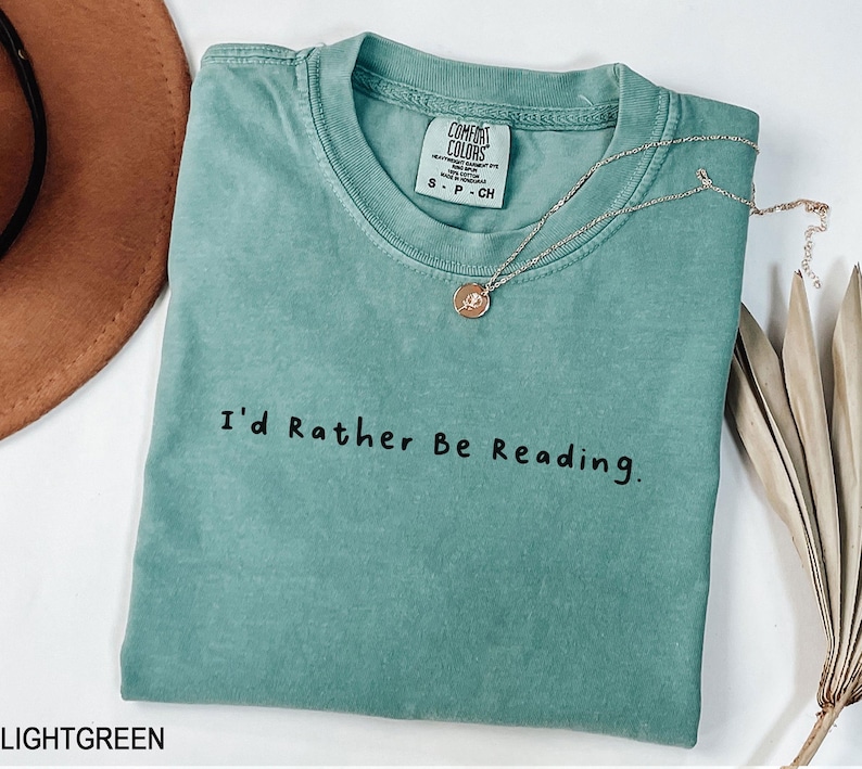 May include: Light green t-shirt with the text "I'd Rather Be Reading." printed in black. The shirt has a distressed look and is made of a soft, comfortable fabric.