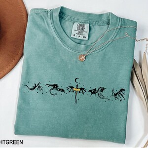 May include: Light green t-shirt with a crew neck and short sleeves. The shirt features a black dragon graphic design across the front. A silver necklace with a pendant is draped across the shirt. The shirt has a tag that says "Comfort Colors".
