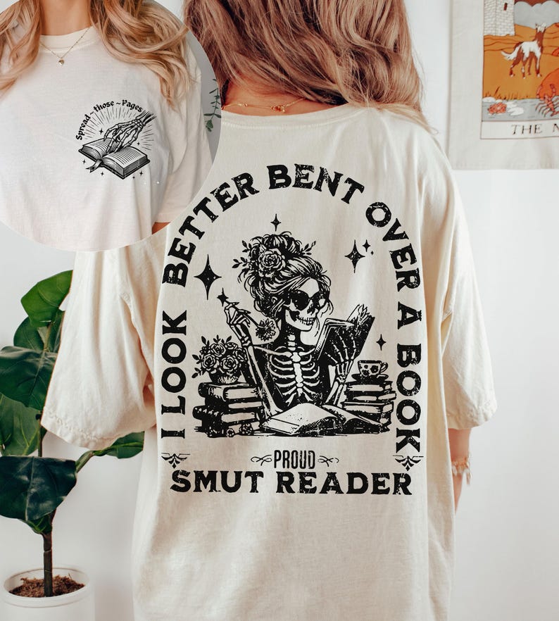 May include: A white t-shirt with a black graphic print of a skeleton woman reading a book. The text on the shirt reads "I look better bent over a book, Proud Smut Reader".