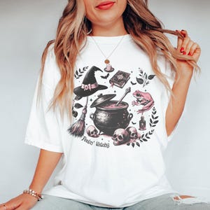 May include: White t-shirt with a pink and black witchy graphic featuring a cauldron, a witch's hat, a frog, a broom, and skulls. The text "Feelin' Witchy" is printed below the graphic.