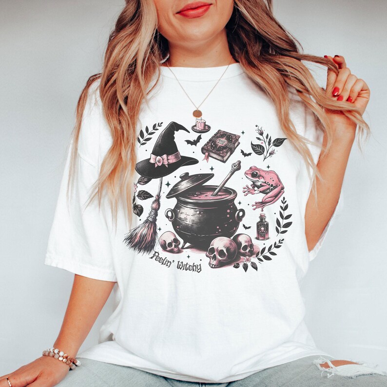 May include: White t-shirt with a pink and black witchy graphic featuring a cauldron, a witch's hat, a frog, a broom, and skulls. The text "Feelin' Witchy" is printed below the graphic.