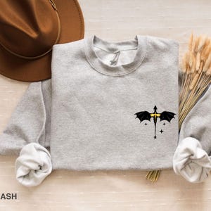 Puede incluir: Una sudadera gris con un gr&aacute;fico de drag&oacute;n negro con detalles amarillos. El drag&oacute;n est&aacute; volando con las alas extendidas. La sudadera est&aacute; sobre una superficie marr&oacute;n claro.