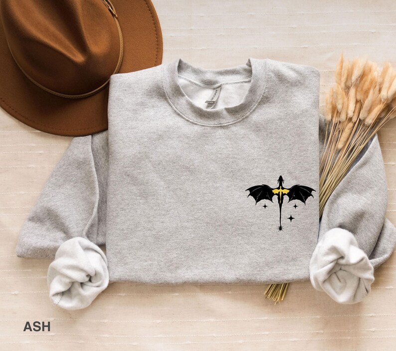 Puede incluir: Una sudadera gris con un gr&aacute;fico de drag&oacute;n negro con detalles amarillos. El drag&oacute;n est&aacute; volando con las alas extendidas. La sudadera est&aacute; sobre una superficie marr&oacute;n claro.
