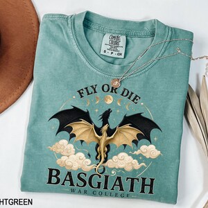 May include: Light green t-shirt featuring a dragon graphic and the text "FLY OR DIE BASGIATH WAR COLLEGE". The dragon has black wings and a gold body. A silver necklace is draped across the shirt.