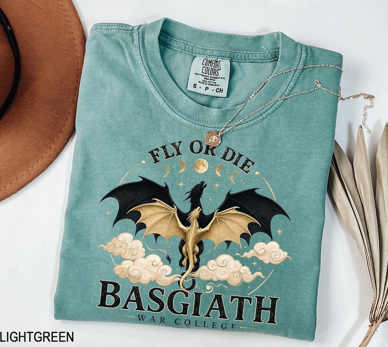 May include: Light green t-shirt featuring a dragon graphic and the text "FLY OR DIE BASGIATH WAR COLLEGE". The dragon has black wings and a gold body. A silver necklace is draped across the shirt.