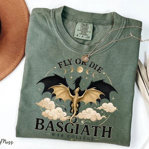 May include: A moss green t-shirt with a dragon graphic. The design features a black dragon with gold wings, surrounded by clouds and the text "FLY OR DIE" and "BASGIATH WAR COLLEGE". A silver necklace rests on the shirt.