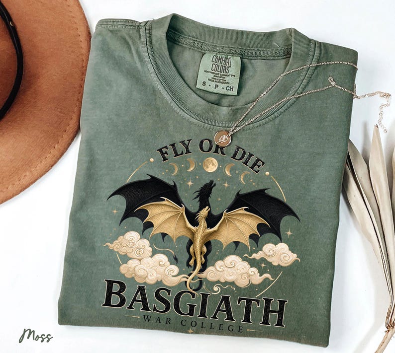 May include: A moss green t-shirt with a dragon graphic. The design features a black dragon with gold wings, surrounded by clouds and the text "FLY OR DIE" and "BASGIATH WAR COLLEGE". A silver necklace rests on the shirt.