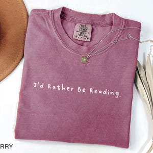 May include: A mauve colored t-shirt with the text "I'd Rather Be Reading." printed in white. The shirt is made of a soft, comfortable material.