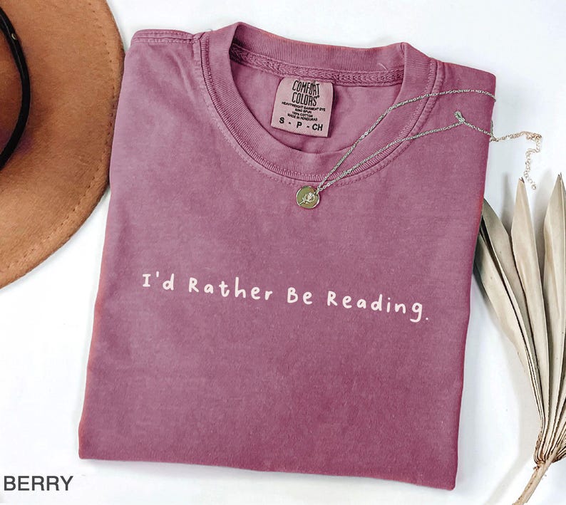 May include: A mauve colored t-shirt with the text "I'd Rather Be Reading." printed in white. The shirt is made of a soft, comfortable material.