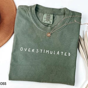 May include: A moss green t-shirt with the text "OVERStimulated" in white lettering. The shirt is made of a soft, comfortable fabric and has a relaxed fit.