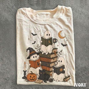 May include: An ivory t-shirt featuring a graphic illustration of three ghosts reading books. One ghost wears a witch hat, while a black cat sits nearby with a pumpkin. The design includes bats, a crescent moon, a candle, and a potion bottle.