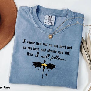 May include: A blue jean colored t-shirt with the text "I chose you not as my next but as my last, and should you fall, then I will follow." A gold necklace and a dragon graphic are also visible.