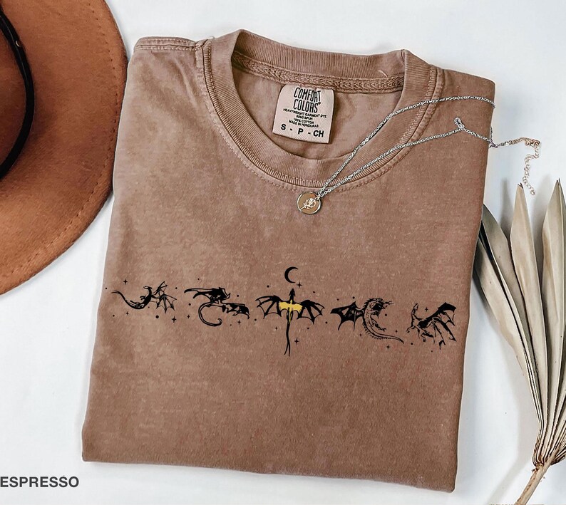 May include: Espresso-colored t-shirt with a crew neck and short sleeves. The shirt features a black dragon graphic design across the chest. A silver necklace with a pendant is draped across the shirt. The shirt has a Comfort Colors tag.